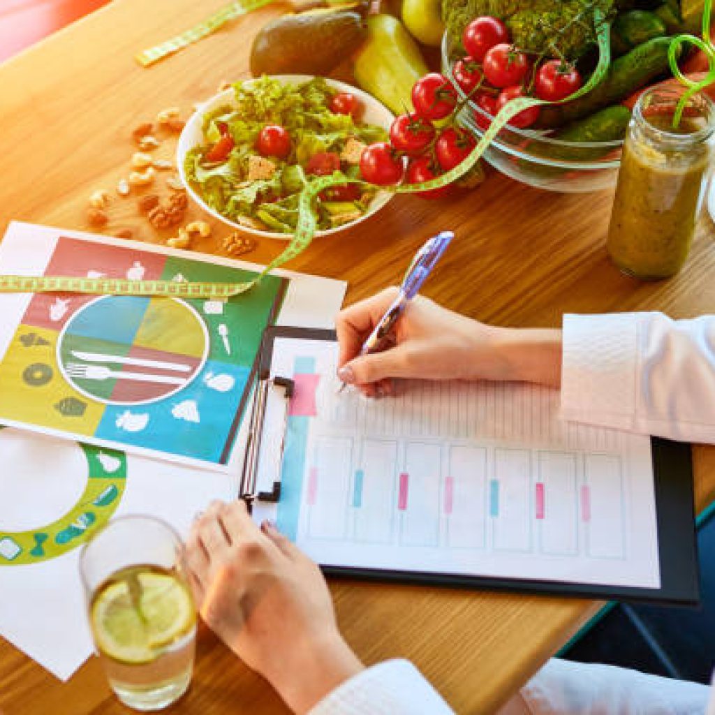 istockphoto-1183411139-612×612 Woman dietitian in medical uniform with tape measure working on a diet plan sitting with different healthy food ingredients in the green office on background. Weight loss and right nutrition concept